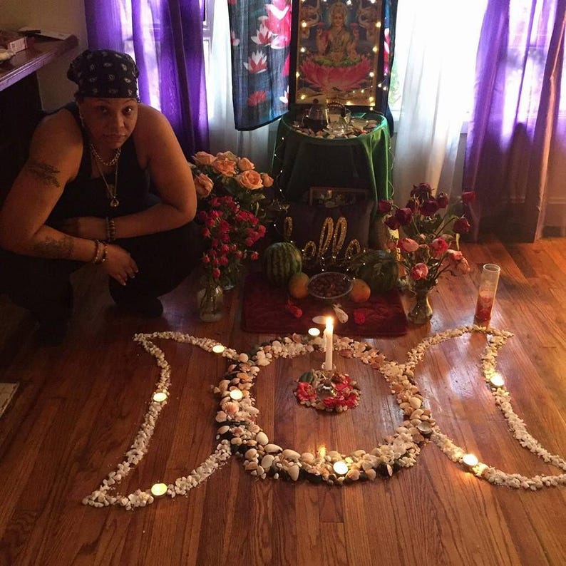 May include: A floor arrangement of white seashells and small stones forming a triple moon symbol on a wooden floor. A central white pillar candle sits within the circle, surrounded by smaller tea light candles placed along the shell outline.