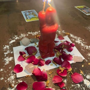 May include: A red candle shaped like two figures embracing, with two lit wicks burning at the top. The candle sits on a white paper with handwritten text, surrounded by scattered dark pink rose petals, light pink crystals, and white powder.