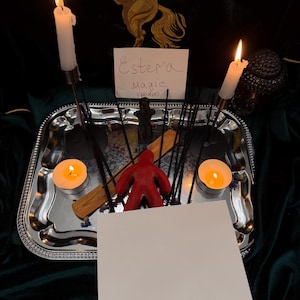 May include: A silver rectangular tray holds two lit white taper candles, two lit tea light candles, several black incense sticks, a red wax figure, and a wooden incense holder. A white card with the handwritten text Estera Magic Voodoo is present.