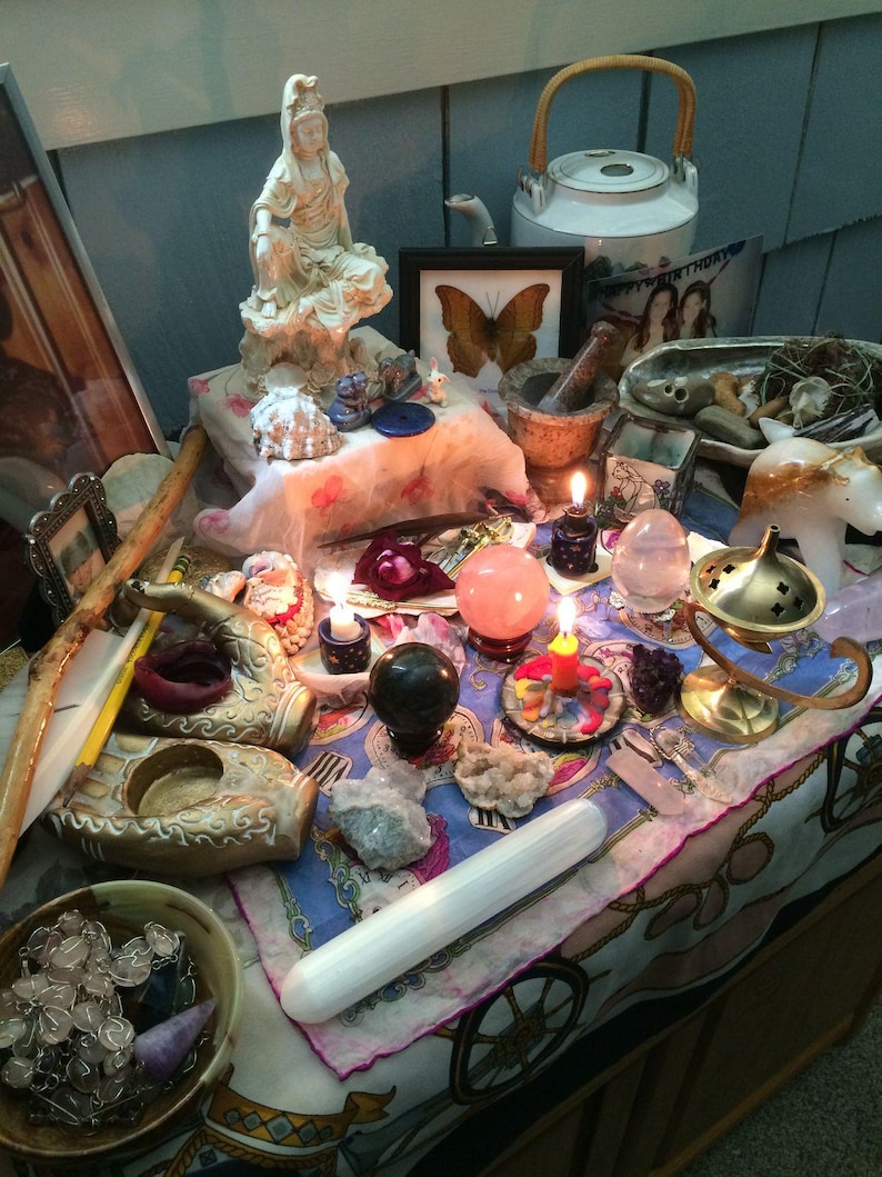 Puede incluir: Una colecci&oacute;n de objetos espirituales sobre un pa&ntilde;o estampado, que incluye una estatua blanca, una tetera, un estampado de mariposa, un mortero, un quemador de incienso de lat&oacute;n, una varita de selenita, varios cristales, esferas y velas encendidas.