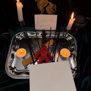 Puede incluir: Una bandeja rectangular plateada contiene dos velas blancas encendidas, dos velas de t&eacute; encendidas, varios palitos de incienso negros, una figura de cera roja y un porta incienso de madera. Una tarjeta blanca muestra el texto escrito a mano Estera Magic Voodoo.
