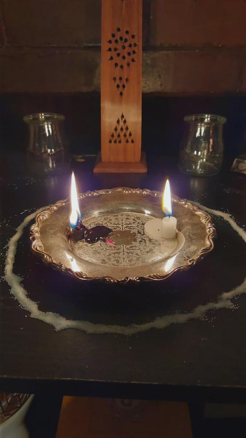 May include: A silver-toned metal plate with an ornate embossed pattern holds two lit candles, one with melting dark wax and one with white wax. A circular ring of white powder surrounds the plate, with a tall wooden incense burner behind it.