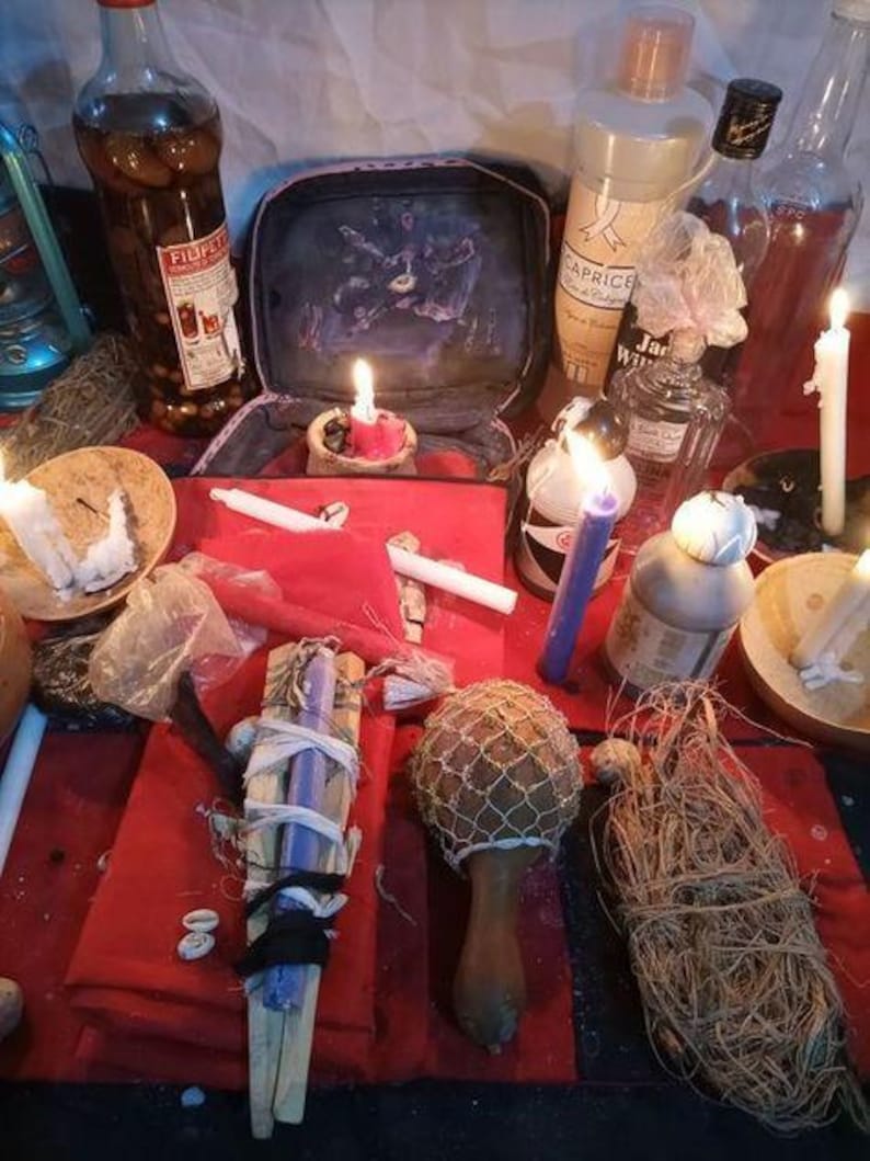 Pu&ograve; includere: Una collezione di oggetti rituali che include un panno rosso, diverse bottiglie di vetro, candele bianche e viola, una maraca in legno con rete, un fascio di fibre essiccate e una piccola custodia aperta con una stampa scura su base rossa.