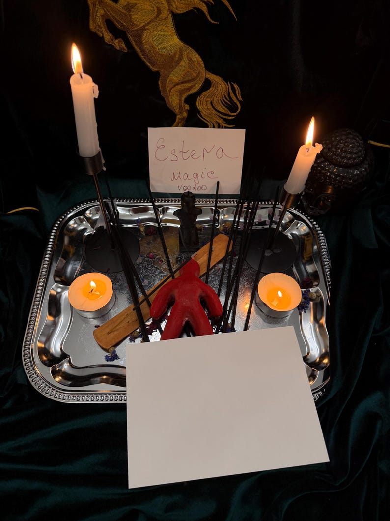 Pu&ograve; includere: Un vassoio rettangolare argentato contiene due candele bianche accese, due lumini accesi, diversi bastoncini di incenso neri, una figura in cera rossa e un porta incenso in legno. Un cartoncino bianco riporta il testo scritto a mano Estera Magic Voodoo.
