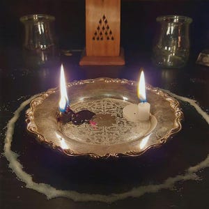 Puede incluir: Un plato de metal plateado con un patr&oacute;n en relieve ornamentado sostiene dos velas encendidas, una con cera oscura derretida y otra con cera blanca. Un anillo circular de polvo blanco rodea el plato, con un quemador de incienso de madera detr&aacute;s.