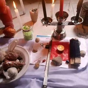 May include: A metal candelabra holding two black candles and one red candle, surrounded by various items including a white bowl filled with shells, a green jar, a long metal rod, a small red paper square, and several lit white candles.