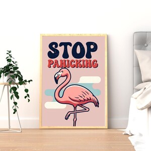 May include: A framed print with a pink flamingo illustration and the text "STOP PANICKING". The flamingo is set against a light pink backdrop with blue and white accents. The frame is a light wood colour. The print is designed to be motivational.