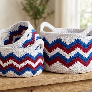 Pode incluir: Três cestos de crochê brancos com padrões de chevron vermelhos e azuis. Os cestos são de tamanhos diferentes e têm alças. Estão em cima de uma mesa de madeira. São uma solução de armazenamento decorativa.