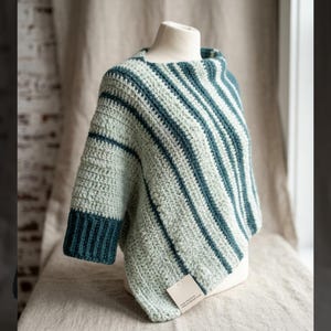 Puede incluir: Poncho de ganchillo hecho a mano en rayas verde claro y verde azulado. El poncho presenta un cuello barco y mangas tres cuartos con puños verde azulado. La prenda se exhibe en un maniquí, mostrando su punto texturizado y su patrón de rayas diagonales.