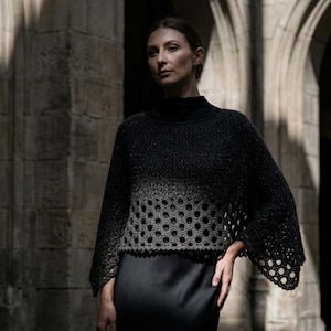 May include: A dark gray crochet top with a unique openwork design. The top features a high neckline and flared sleeves. The openwork pattern transitions from a solid dark gray at the top to a lighter gray at the bottom, creating a gradient effect. The top is paired with a black skirt.