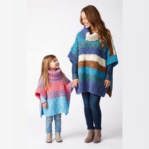May include: A woman and a child wearing matching, colourful, striped ponchos with turtleneck collars. The woman's poncho is blue, white, and brown. The child's poncho is pink, purple, and blue. Both are holding hands.