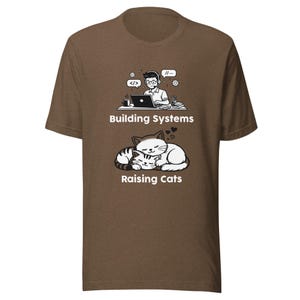 Op de afbeelding: Bruin t-shirt met een afbeelding van een programmeur achter een laptop boven twee cartoonkatten. De tekst luidt "Building Systems" en "Raising Cats". Het ontwerp is in zwart en wit.