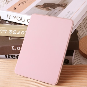 May include: A light pink tablet case rests on a wooden surface, with the text "IF YOU CAN" visible on a book in the background. Other books are stacked nearby, creating a layered composition. The scene is lit with soft, natural light.