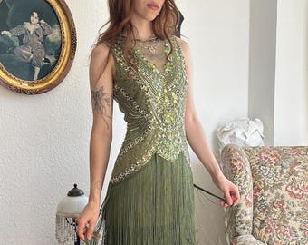 Puede incluir: Un vestido sin mangas de color verde oliva con intrincados abalorios y lentejuelas en un patrón de cuello en V, con múltiples capas de largos flecos que se extienden desde la cintura hasta el dobladillo. Un tocado negro con plumas y cuentas oscuras.