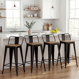 May include: Four black metal bar stools with wooden seats are arranged in a row. The stools have a simple design with a backrest and footrest. The kitchen setting includes a white countertop, white subway tile backsplash, and natural wood flooring.