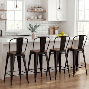May include: Four black metal bar stools with wooden seats are arranged in a row. The stools have a curved backrest and a footrest. The kitchen setting includes a white countertop, a white brick backsplash, and a window.