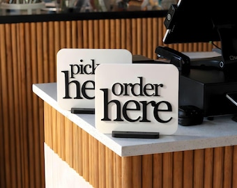 May include: Two white rectangular signs with rounded corners, each with black text. One says "pick here" and the other says "order here." The signs are on black stands, placed on a white countertop.