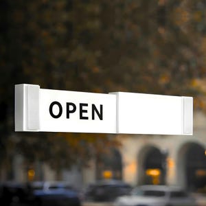 May include: A white rectangular sign with the word "OPEN" in bold black letters. The sign is mounted on a glass door, with a blurred outdoor scene visible through the glass.