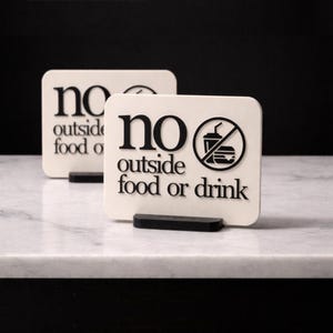 May include: Two cream-colored signs with the words "no outside food or drink" in black. One sign has a graphic of a drink and burger crossed out. The signs are on black stands, set on a white marble surface.