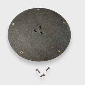 Op de afbeelding: Een donkergrijs, cirkelvormig object met een getextureerd oppervlak, met verschillende kleine gaatjes en messingkleurige klinknagels rond de rand. Drie kleine schroeven zijn eronder geplaatst.