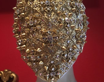 Può includere: Una maschera dorata con un design intricato e strass trasparenti. La maschera è ricoperta di punte dorate.