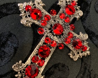 May include: A silver cross pendant necklace with red crystal embellishments. The cross is ornate and features a detailed design.