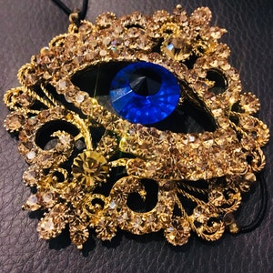 May include: A gold-tone evil eye charm with a blue eye and clear rhinestones. The charm is attached to a black string.