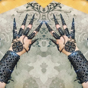 May include: A pair of black metal gloves with intricate designs and black gemstones. The gloves have long, pointed fingers that resemble claws.