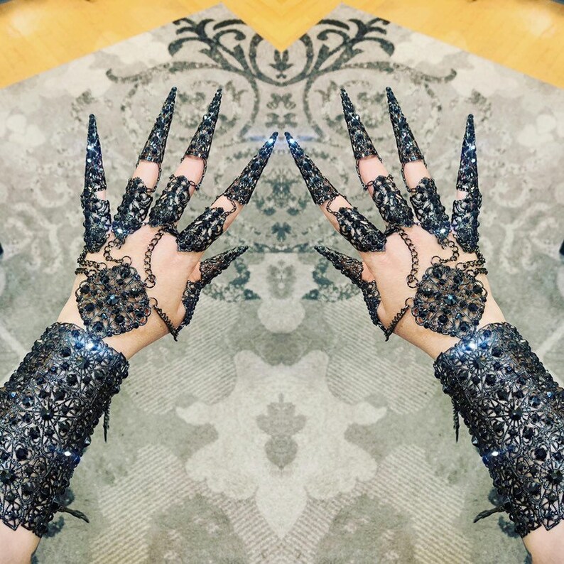 May include: A pair of black metal gloves with intricate designs and black gemstones. The gloves have long, pointed fingers that resemble claws.