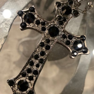 May include: A silver cross pendant with black rhinestone accents. The cross is detailed with a filigree design and has a chain attached.