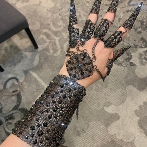 May include: A black metal glove with a chain and a large, ornate design. The glove has long, pointed fingers and is covered in rhinestones.