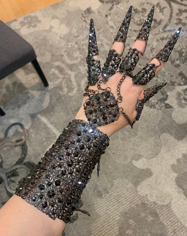 May include: A black metal glove with a chain and a large, ornate design. The glove has long, pointed fingers and is covered in rhinestones.