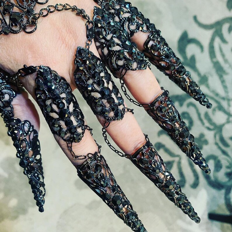 May include: Black metal finger rings with intricate designs and rhinestones. The rings are connected by chains and extend into long, pointed claws.