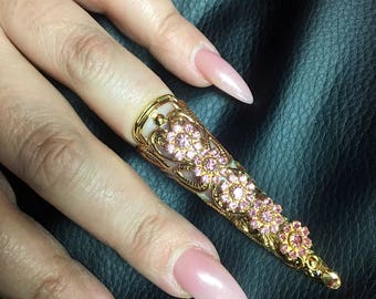 May include: A gold-colored finger nail ring with pink gemstones in a floral design. The ring covers the entire nail and is worn on a finger with a long, pointed, pink nail. The background is black.