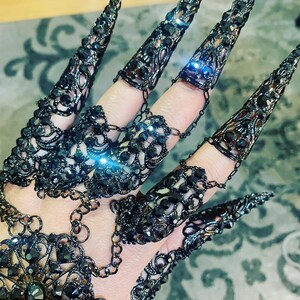 May include: A set of black metal claw rings with rhinestones. The rings are connected by chains and can be worn on multiple fingers.