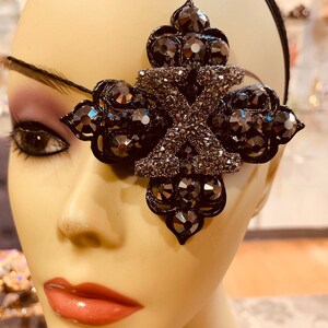 May include: A black eye patch with a large, ornate, silver-toned, crystal-encrusted design. The design features a large, stylized letter "X" in the center.