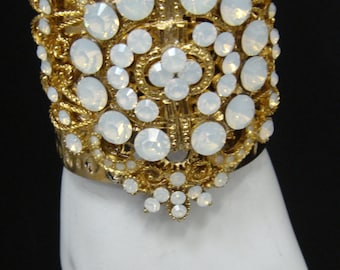 May include: Gold bracelet with a large, ornate design featuring white rhinestones. The bracelet is on a white mannequin hand.