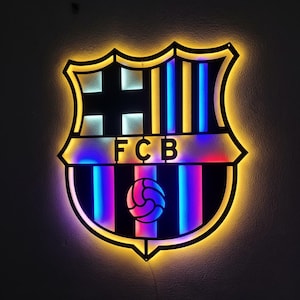 Puede incluir: Logotipo iluminado del FC Barcelona con un contorno negro y colores neón vibrantes. El diseño presenta el escudo del equipo, incluyendo las letras "FCB" y un balón de fútbol. El logotipo está iluminado con luces azules, rosas y amarillas.