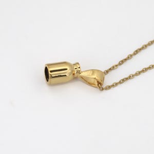 Può includere: Una collana con pendente color oro con un piccolo ciondolo a forma di bottiglia. Il ciondolo ha un corpo cilindrico e una parte superiore arrotondata, con una piccola apertura all'estremità. La catena della collana è delicata e anch'essa color oro.