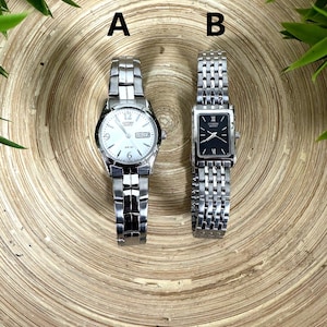 Könnte beinhalten: Zwei silberfarbene Armbanduhren werden gezeigt. Uhr A hat ein rundes weißes Zifferblatt mit schwarzen Ziffern und einer Tages-/Datumsanzeige. Uhr B hat ein rechteckiges schwarzes Zifferblatt mit silbernen Zeigern und römischen Ziffern. Beide Uhren haben silberne Gliederarmbänder.