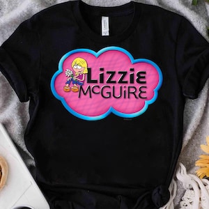 May include: A black short-sleeved t-shirt featuring a graphic of a cartoon character holding a white flower. The design is set within a pink cloud shape with a blue outline and includes the black text Lizzie McGuire printed on the front.