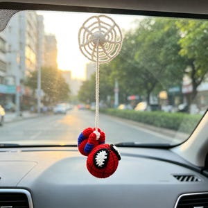 May include: A crocheted car rearview mirror hanging ornament featuring a white circular spider web connected by a long string to a small, round, red and blue character head with large white eyes and black outlines, crafted from yarn.