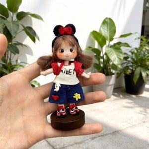 May include: A handmade crochet doll with brown hair, wearing a black headband with a red bow, a white shirt with "Mikey" on it, a red jacket, blue shorts, and red shoes. The doll is standing on a small wooden base.