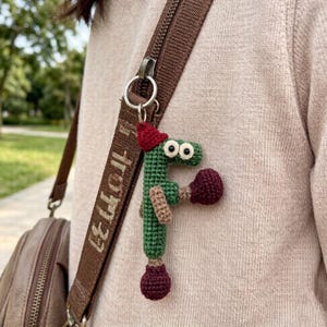 May include: A green crocheted letter "F" keychain with a red hat and boxing gloves. The keychain is attached to a brown strap with white text. The letter has black eyes and is wearing a red hat and boxing gloves.