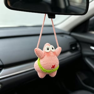 May include: A crochet hanging ornament shaped like a pink starfish character with large white eyes, a lime green waistband, and a small red detail on the belly, suspended by a pink yarn loop from a vehicle rearview mirror.