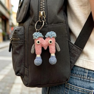 May include: A handmade crochet keychain in the shape of a pink letter "M" with blue accents. The keychain has two small black eyes and gray arms. It is attached to a black backpack with a silver zipper.