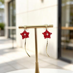 May include: A pair of red flower earrings with a pearl center and a gold-colored, curved dangle. The earrings are displayed on a gold-colored earring stand. The earrings are a floral design.