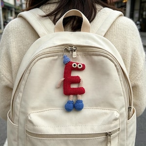 May include: A red crocheted letter "E" keychain with a blue hat and blue shoes, attached to a cream-colored backpack. The letter has googly eyes and tan arms. The backpack has a front pocket and a zipper closure.