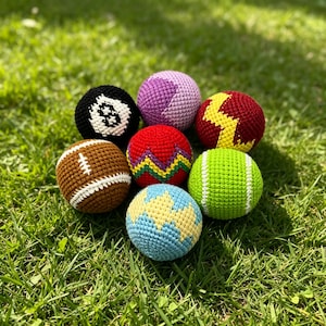May include: A collection of seven spherical crochet balls featuring various designs including a black eight ball, a brown football, a red ball with a yellow zigzag, a light blue ball with yellow patterns, a lime green ball, and a purple ball.