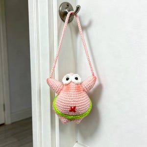 May include: A handmade crochet bag shaped like a pink starfish character with large white eyes, black pupils, and a lime green waistband. The bag features a small red cross detail on the front and includes a long, matching pink crochet strap.